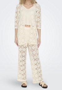 Lace-knit cream cardigan and matching wide-leg pants with geometric patterns, featuring a cropped beige top and flat sandals.