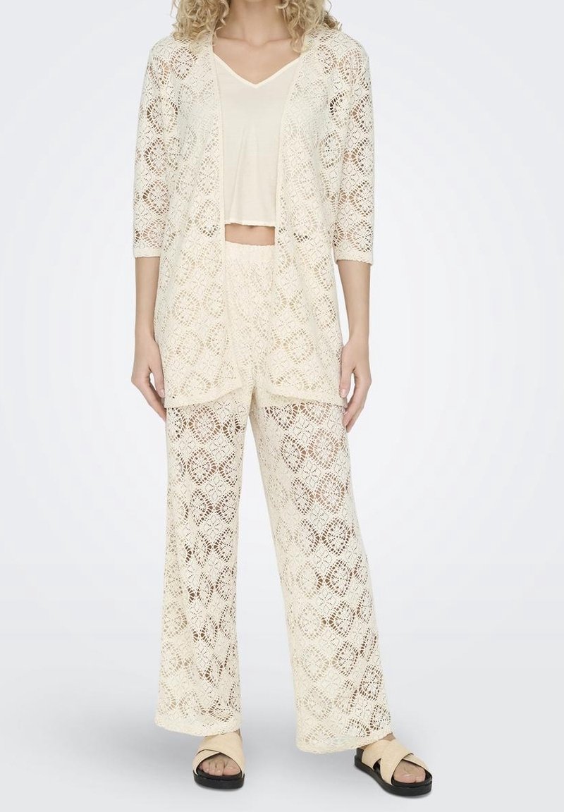 Lace-knit cream cardigan and matching wide-leg pants with geometric patterns, featuring a cropped beige top and flat sandals.