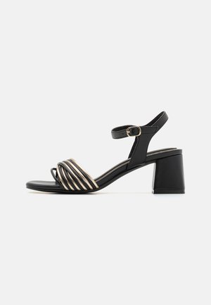 Sandales noires à talons avec un talon bloc et des détails de brides métalliques croisées. Elles disposent d'une bride de cheville réglable et d'une texture lisse.