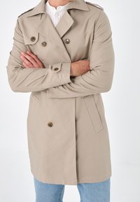 Cappotto beige con bottoni doppi, spalline, tasche laterali e taglio dritto. Indossato sopra jeans di denim.