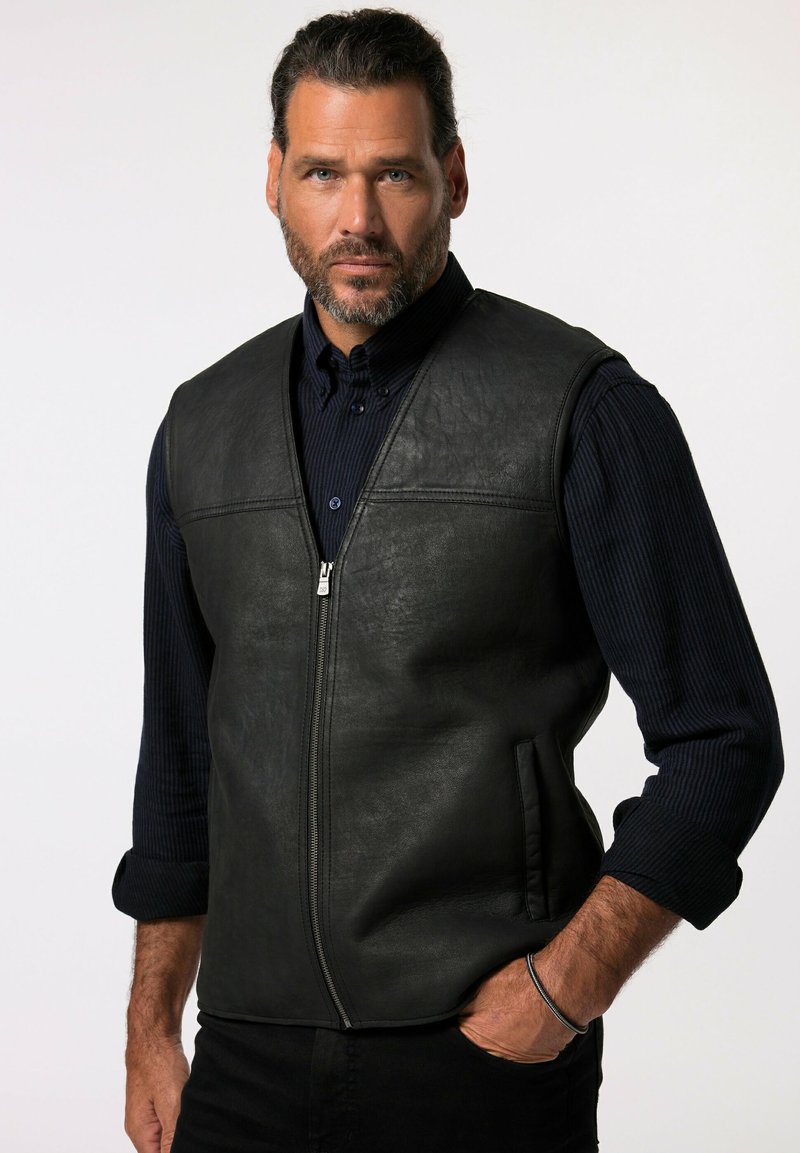 Schwarze Lederweste mit Reißverschluss vorn, zwei Seitentaschen und einer glatten Textur. Getragen über einem dunklen, gerippten Langarmshirt.