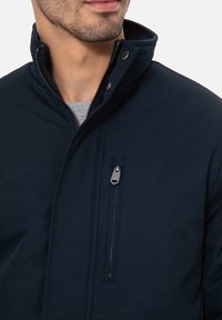 Giacca blu navy con colletto alto, chiusura con zip e tasca sul petto. Il tessuto sembra liscio e resistente, con dettagli in metallo.