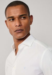 White button-up shirt with a classic collar, featuring a small light blue logo on the chest, smooth texture, and fitted design.