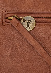 Braune Ledertasche mit Reißverschluss, genähten Kanten, einem Reißverschluss aus Messing und einem runden Messinganhänger mit einem gravierten abstrakten Logo.
