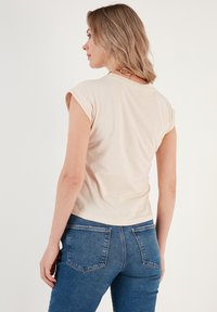 Top beige a maniche corte realizzato in morbido tessuto, con una vestibilità comoda e risvolti sui polsini. Abbinato a jeans blu aderenti.