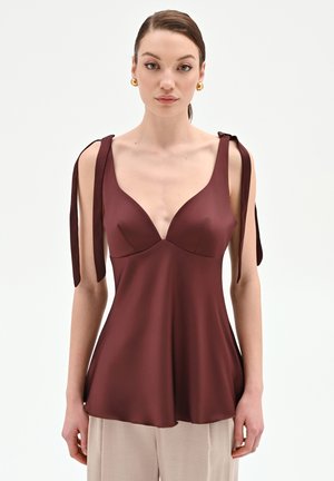 Donna con top senza maniche in colore borgogna scuro con spalline annodate e pantaloni beige, in piedi davanti a uno sfondo bianco semplice.