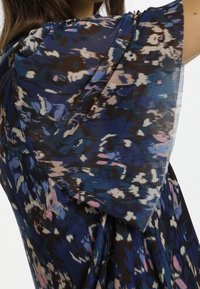 Průhledná blůza v modré, hnědé a růžové abstraktní vzoru. Má krátké, široké rukávy a splývavý design s lehkým materiálem.