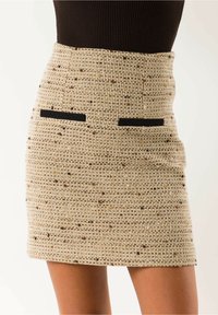 Minifalda de tweed beige de talle alto con motas marrones y dos detalles horizontales de bolsillos negros, combinada con una blusa oscura.