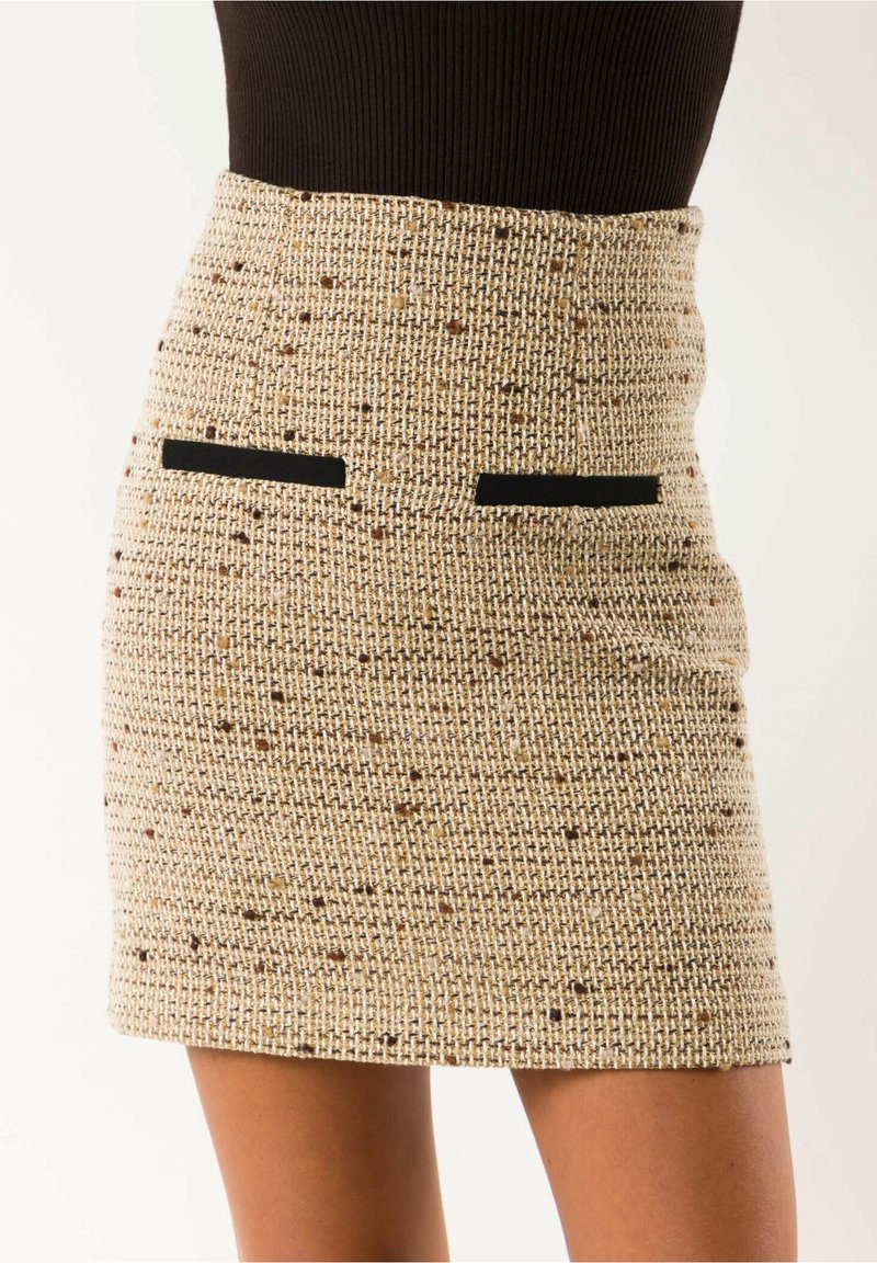 Minifalda de tweed beige de talle alto con motas marrones y dos detalles horizontales de bolsillos negros, combinada con una blusa oscura.