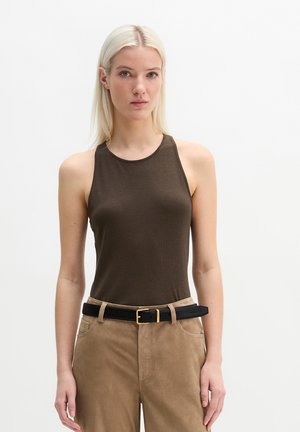 Jeune femme aux cheveux blonds raides, portant un haut marron foncé sans manches, un pantalon beige et une ceinture noire à boucle dorée.