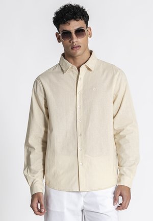 Junger Mann, der ein beiges, langärmeliges Hemd mit Knöpfen, weiße Hosen und dunkle Sonnenbrillen trägt, steht vor einem schlichten weißen Hintergrund.