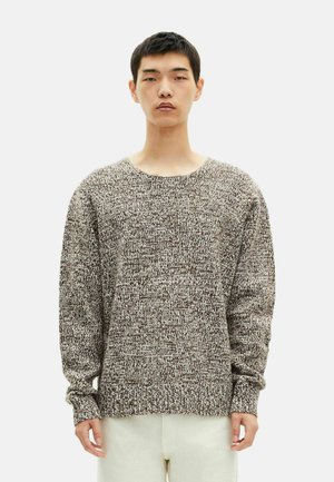 Brun og creme strikket sweater med bred udsnit, lange ærmer og et struktureret mønster. Kombineret med lyse bukser.