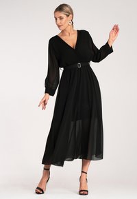 Czarna maxi sukienka z głębokim dekoltem w kształcie V, plisowanym topem, długimi przezroczystymi rękawami, wcięciem w talii z klamrą oraz warstwową, zwiewną spódnicą.