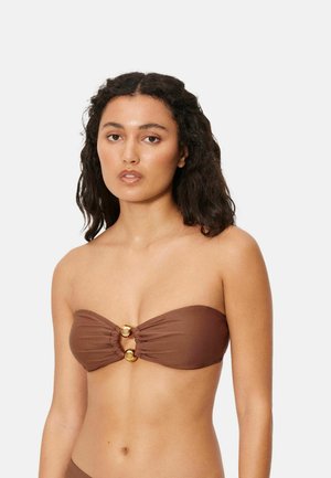 Mujer con cabello oscuro y ondulado, vestida con un top de bikini marrón sin tirantes con dos detalles de anillos dorados en el centro, sobre un fondo blanco.