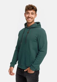 Grüner Hoodie aus strukturiertem Stoff, mit Kängurutasche, gerippten Bündchen und einem Kapuzenzug mit schwarzen und weißen Akzenten.