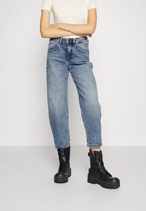Personne portant un jean bleu clair taille haute et des bottes noires épaisses à lacets, bras croisés devant un fond blanc uni.