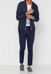 Veste légère bleu marine avec fermeture éclair à l'avant et accents blancs, associée à des baskets grises, un pantalon bleu marine et un polo blanc avec un col orange.