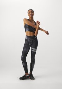 Sportovní podprsenka a legíny s černobílým batikovaným vzorem a stříbrnými akcenty. Modelka má na sobě černé sportovní boty a natahuje se s jednou rukou.