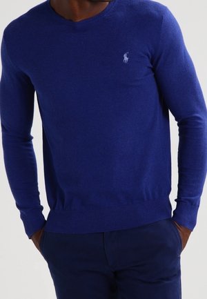 Mann trägt einen eng anliegenden blauen V-Ausschnitt-Pullover mit einem kleinen weißen, auf der Brust gestickten Logo, Hände in den Taschen einer dunkelblauen Hose.