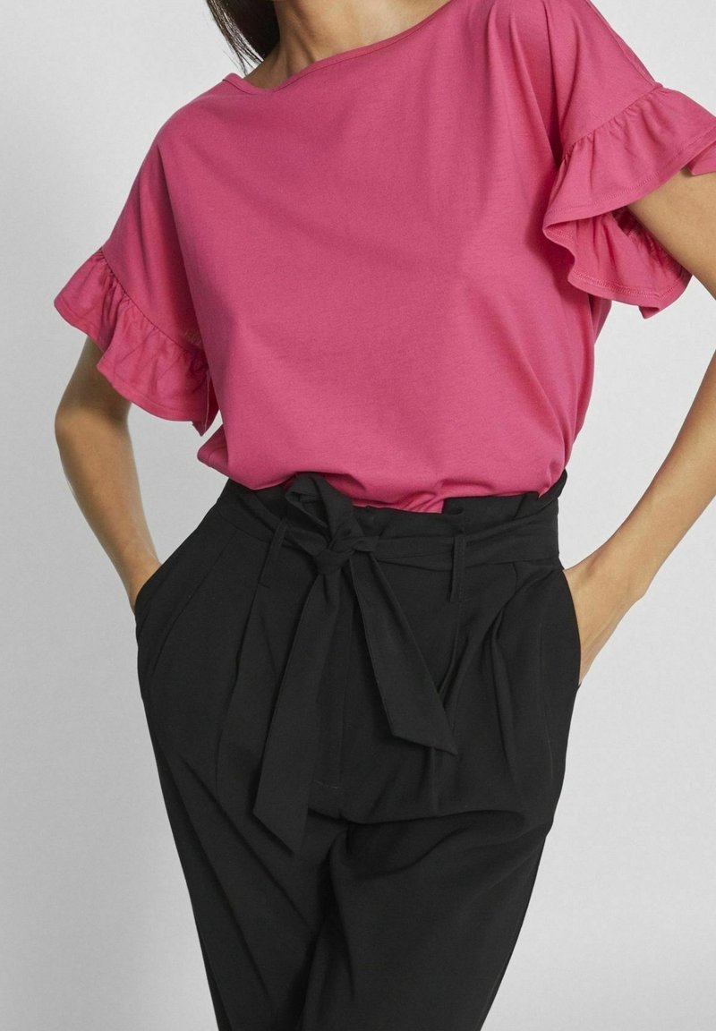 Femme portant un haut rose à manches courtes à volants et un pantalon paperbag noir taille haute noué avec une ceinture en tissu.