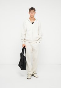 Felpa con zip di colore beige chiaro e pantaloni abbinati, realizzati in tessuto morbido. Borsa a mano nera, abbinata a sneaker off-white con dettagli neri.