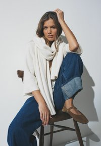 Hvid strikket sweater, oversized tørklæde og brede blå jeans. Brun ruskind flats med stropdetalje. Model i en siddende position på en træstol.