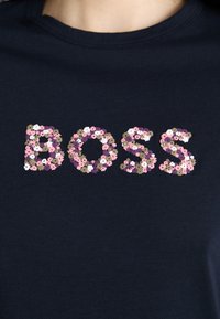 Námořnická košile s nápisem "BOSS" vyvedeným v malých květinových vzorech v růžové, bílé, fialové a hnědé barvě na hrudi.