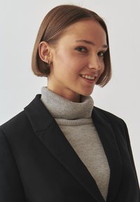 Mustamantel halli kõrvarõngaga ja hall turtlenecki kampsuniga. Lühikesed sirged pruunid juuksed, minimaalsed kuldse väli kõrvarõngad, neutraalne taust.