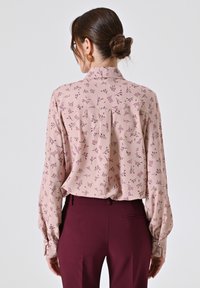 Blusa rosa chiaro con motivo floreale, maniche lunghe e colletto alto, abbinata a pantaloni borgogna su misura. Tessuto dalla trama liscia.