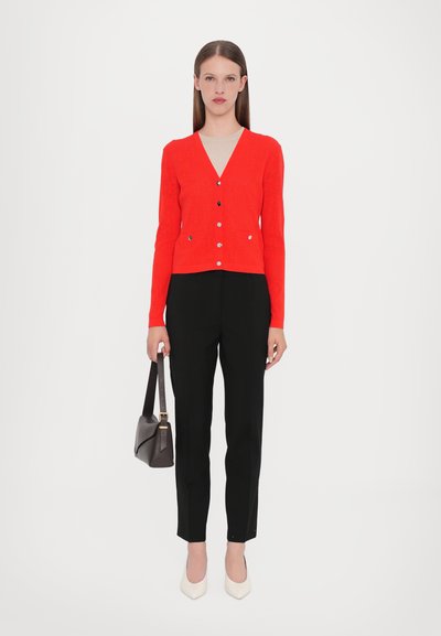 Cardigan rosso con scollo a V e maniche lunghe, con chiusure a bottone e tasche, abbinato a pantaloni neri su misura e tacchi bianchi.
