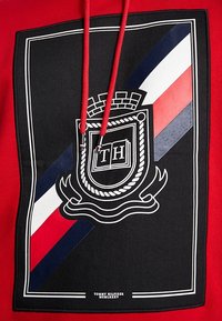 Sudadera roja con un parche negro que presenta un emblema de cuerda blanca, rayas diagonales en rojo, blanco y azul marino, y las iniciales "TH" en un diseño de libro abierto.