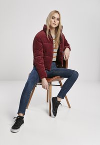 Burgunderfarget kordfløyelsjakke, grå og gulstripete skjorte, blå skinny jeans, svarte sneakers, sittende på en trestol mot en ensfarget bakgrunn.