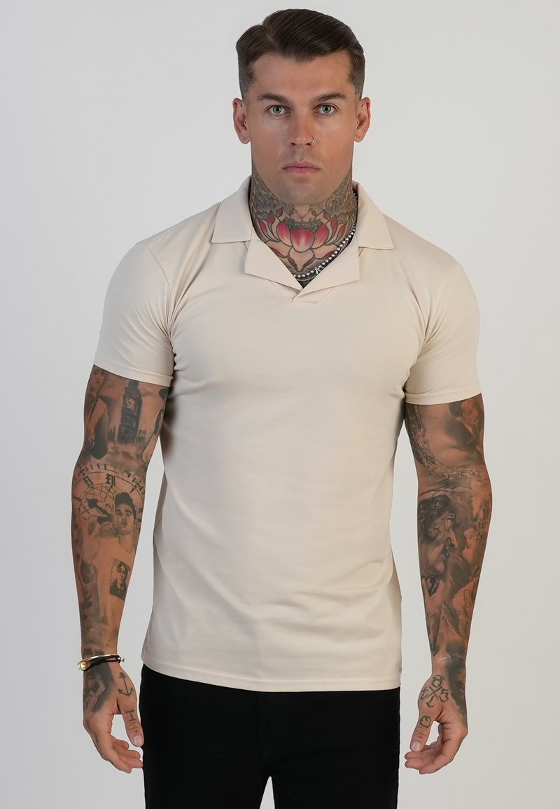 Beige polo shirt met korte mouwen, een v-hals en een zachte textuur, gedragen door een model met tatoeages, tegen een effen achtergrond.