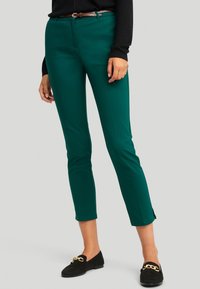 Zelené kalhoty po kotníky s hladkou texturou, zapínáním na zip a knoflík, bočními rozparky a slim fit. V kombinaci s černými mokasíny.