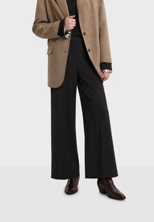 Personne portant un pantalon noir à jambes larges, un blazer marron sur un top noir avec des poignets ornés, et des bottines à talons brun foncé.