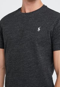 Mörkgrå t-shirt med kort ärm i strukturerad tyg, som har en liten vit logotyp på vänster bröst. Rund halsringning.