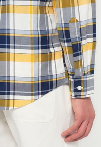Männliche Hand neben der Hüfte in weißen Hosen, die ein kariertes Hemd in Gelb, Blau, Grau und Weiß mit geknöpften Bündchen und abgerundetem Saum trägt.