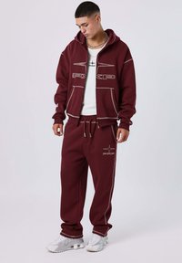 Conjunto de sudadera y pantalones de chándal burdeos, con costuras blancas y detalles de logo. La sudadera cuenta con un bolsillo en la parte delantera; los pantalones tienen una cintura elástica.