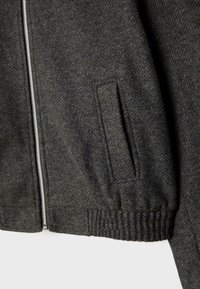 Section de veste en laine gris foncé montrant une fermeture éclair frontale, une poche latérale avec rabat et un ourlet côtelé élastique.