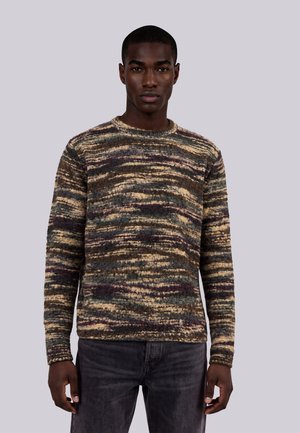 Uomo che indossa un maglione a maniche lunghe con trama e righe orizzontali in tonalità della terra e pantaloni grigio scuro, in piedi davanti a uno sfondo semplice.
