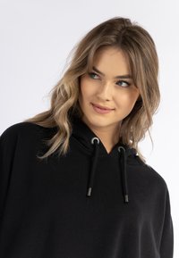 Tânără cu păr blond ondulat, purtând un hanorac negru, privind în lateral cu un zâmbet ușor pe fundal alb.