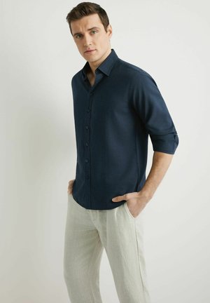 Mężczyzna w ciemnoniebieskiej koszuli z guzikami z podwiniętymi rękawami oraz jasnobeżowych spodniach, stoi z rękami w kieszeniach na jednolitym tle.