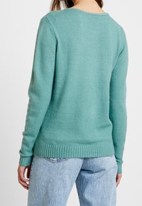 Ljust turkosfärgad tröja med rund halsringning, ribbade muddar och nederkant, tillverkad av mjukt material. Bärs med ljusblå denimjeans, sedd bakifrån.