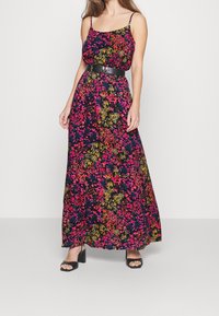 Maxiklänning med tunna spaghetti-remmar, svart tyg täckt av färgglada blommönster, åtdragen vid midjan med ett svart bälte. Svarta högklackade sandaler.