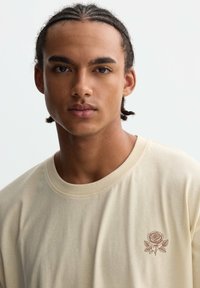 Camiseta de color crema con mangas cortas, que presenta un pequeño bordado de una rosa marrón en el lado izquierdo del pecho. Textura de tela suave y lisa.