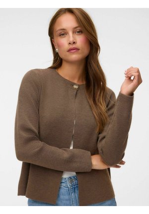 Femme aux longs cheveux châtain clair portant un cardigan marron à un bouton sur un haut blanc et un jean bleu, posant avec un bras levé.