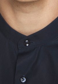 Donkerblauw overhemd met knopenlijst, voorzien van een afgeronde kraag, een gladde stoftextuur en twee zichtbare knopen bij de halslijn.