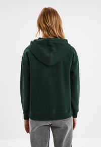 Dunkelgrüne Kapuzenjacke mit Zugkordel, gerippten Bündchen und Saum, aus weichem Material. Die Rückansicht zeigt einen lässigen Schnitt und keine sichtbaren Muster.
