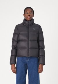 Black puffer jacket with a high collar, zip closure, and horizontal seam detailing. Features logo on the left chest and a smooth finish.