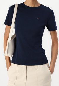 Navy cotton T-shirt with short sleeves and crew neckline, featuring a small red and white logo on the chest. Worn with light beige trousers.